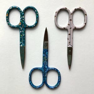May include: Three pairs of small, decorative scissors with curved handles. One pair has a teal background with a floral pattern, another has a light pink background with a small insect pattern, and the third has a blue background with a floral pattern.