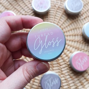 May include: A hand holds a small, round, silver tin of "Thread Gloss." The lid features a pastel gradient of pink, blue, and yellow with the words "Spring Awakening" and "Thread Gloss" in white script. Other tins with pink and purple lids are scattered around.