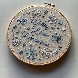 May include: Embroidered hoop art with the phrase "Walking in a Winter Wonderland" in blue thread. The design features snowflakes in various sizes and shades of blue, set against a neutral fabric background, and framed by a wooden hoop.
