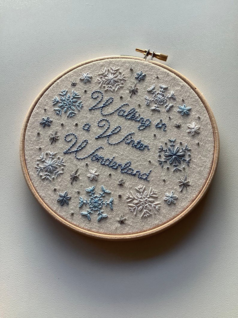 May include: Embroidered hoop art with the phrase "Walking in a Winter Wonderland" in blue thread. The design features snowflakes in various sizes and shades of blue, set against a neutral fabric background, and framed by a wooden hoop.