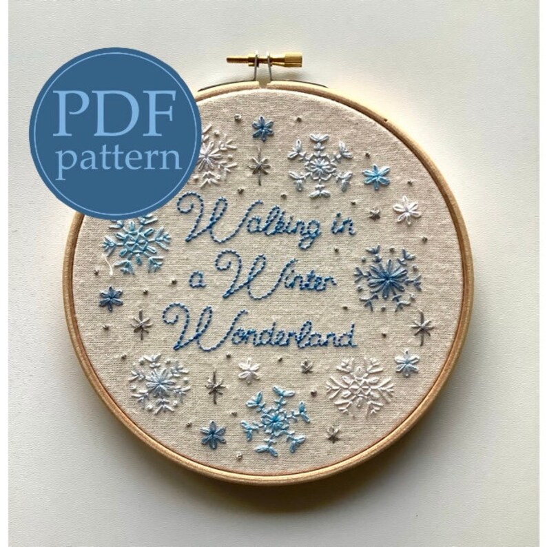 May include: An embroidery hoop featuring a winter theme. The design includes the phrase "Walking in a Winter Wonderland" in blue thread, surrounded by embroidered snowflakes in shades of blue and white. A blue circle with "PDF pattern" is in the upper left.