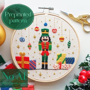 May include: An embroidery hoop with a preprinted Christmas nutcracker design. The design features a nutcracker in a green and red uniform, hanging ornaments, and wrapped gifts. The text "Preprinted pattern" is in a green circle.