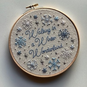 May include: Embroidered hoop art with the phrase "Walking in a Winter Wonderland" in blue script. The design features snowflakes in shades of blue and white, set against a neutral fabric background, all within a wooden hoop.