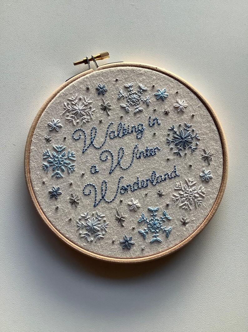 May include: Embroidered hoop art with the phrase "Walking in a Winter Wonderland" in blue script. The design features snowflakes in shades of blue and white, set against a neutral fabric background, all within a wooden hoop.