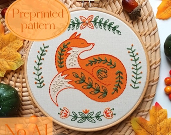 May include: Embroidered fox preprinted pattern in a wooden hoop. The fox is orange with white accents and surrounded by green and orange floral and leaf designs. The text "Preprinted pattern" is in an orange oval. The text "No AI. 100% human-made" is in an orange rectangle.