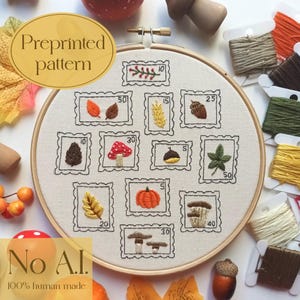May include: An embroidery hoop containing a cream-colored fabric with a preprinted pattern of twelve rectangular postage stamp designs. Each stamp features embroidered autumn motifs including leaves, mushrooms, acorns, and a pumpkin, with numerical values.
