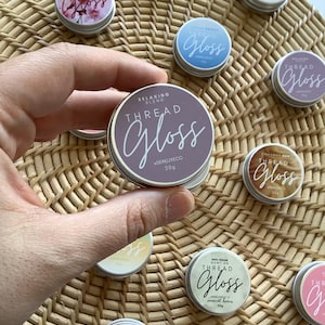 May include: A collection of small, round tins of Thread Gloss, with the main tin held in a hand. The tins have a metallic silver base and lids with the words "Thread Gloss" in a script font. The tins are in various colors.