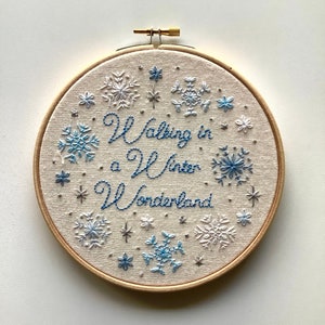 May include: Embroidered hoop art with the phrase "Walking in a Winter Wonderland" in blue thread. The design features snowflakes in shades of blue and white, set against a beige fabric background, and framed by a wooden hoop.