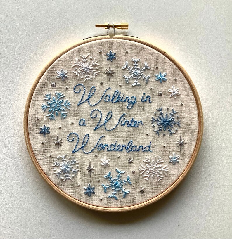 May include: Embroidered hoop art with the phrase "Walking in a Winter Wonderland" in blue thread. The design features snowflakes in shades of blue and white, set against a beige fabric background, and framed by a wooden hoop.