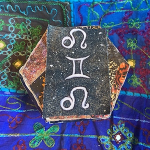 Pode incluir: Uma pilha de pequenos quadrados de tecido preto e branco com símbolos astrológicos. Os símbolos são Leão e Gêmeos. Os quadrados de tecido estão sobre um fundo de tecido colorido e estampado.