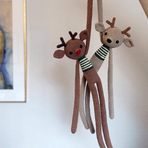 May include: Two handmade crochet reindeer toys. One reindeer is brown with a red nose and a green and white striped scarf. The other reindeer is tan with a green and white striped scarf. Both have long legs and antlers.