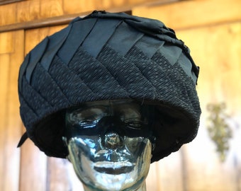 May include: A black, pleated fabric hat with a wide brim. The hat is on a mannequin head.