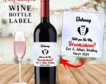 May include: A personalized wine bottle label with a tuxedo graphic and the text "Will you be my Groomsman? Lori & Adam's Wedding March 20,24". The label is white with black text and a black tuxedo graphic.