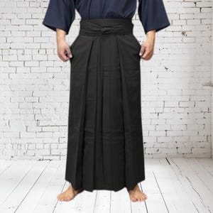 May include: A black hakama, a traditional Japanese garment, is shown. The hakama is pleated and worn with a dark blue top. The garment is tied at the waist with a knot. The person is standing in front of a white brick wall.