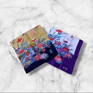 以下が含まれることがあります： 複雑な花柄が描かれた2枚の正方形の布製ハンカチ。1枚はゴールド地に濃紺の縁取り、もう1枚は白地に紫の縁取りです。どちらも鮮やかな赤、青、紫の花が描かれており、日本語の文字がプリントされています。