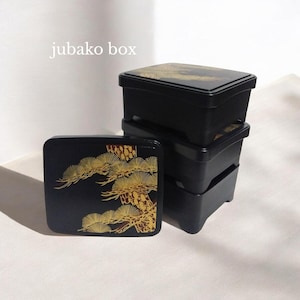 Peut inclure: Une pile de trois boîtes rectangulaires noires de type jubako avec un couvercle placé devant. Les couvercles présentent un motif de pin doré. Le texte "jubako box" est imprimé en blanc dans le coin supérieur gauche de l'image.
