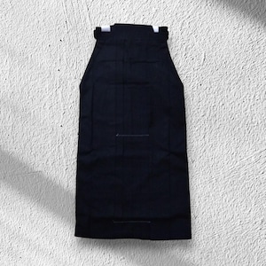 May include: A black hakama, a traditional Japanese garment, is displayed against a textured white background. The hakama features vertical pleats and a rectangular shape, with a structured waistband and a simple design.
