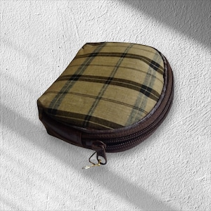 May include: A small, half-moon-shaped pouch with a brown zipper and a plaid pattern in shades of tan, brown, and black. The pouch has a brown leather-like base and a metal zipper pull.