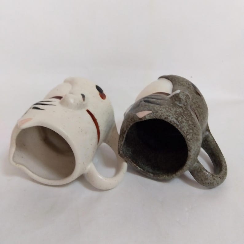 May include: Two ceramic mugs with cat faces. One mug is white with black and brown stripes and the other is gray with black stripes.