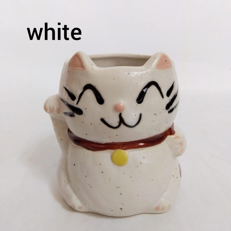 May include: White ceramic cat planter with black eyes and a brown collar. The cat is smiling and has a yellow bell on its collar.