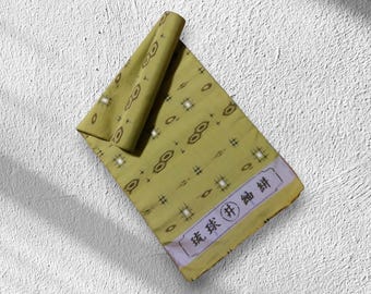 May include: A folded, olive-green textile with a repeating pattern of eye-like shapes and hash marks. The fabric has a rolled section at the top. The bottom edge features a rectangular label with Japanese text.