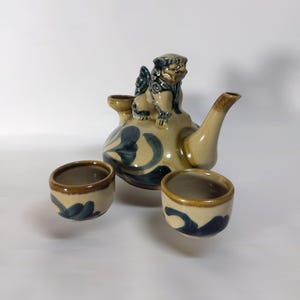 以下が含まれることがあります： ベージュの釉薬に濃い青色の花柄が描かれた陶器製の急須で、蓋にはライオンの彫刻が施されています。急須には湾曲した注ぎ口があり、同じ青い花柄が描かれた小さな陶器製の湯呑みが2つセットになっています。