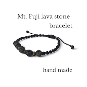 Puede incluir: Una pulsera de cordón negro con cuatro piedras de lava porosas de color gris oscuro del Monte Fuji, separadas por pequeños espaciadores de metal dorado. La pulsera tiene un cierre de nudo deslizante y el texto Mt. Fuji lava stone bracelet hand made.