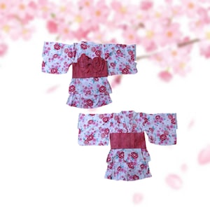 以下が含まれることがあります： 伝統的な日本の夏の着物である、花柄の浴衣が2着展示されています。浴衣は、ピンクと赤の花柄の白い背景が特徴です。それぞれに赤い帯とティアードスカートが付いています。背景には、ピンクの桜がぼやけています。