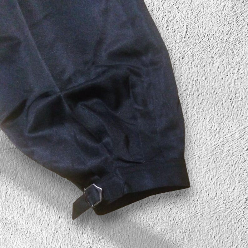 May include: A close-up shot of dark navy fabric, possibly a sleeve or trouser leg, featuring a small silver buckle and strap. The material has a smooth, slightly glossy appearance, with visible folds and creases.