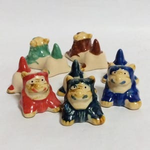 以下が含まれることがあります： 赤、青、緑、茶色、白など、さまざまな色の猫のセラミック製置物7個セット。猫は白いセラミック製の小さな塚の上に座っています。