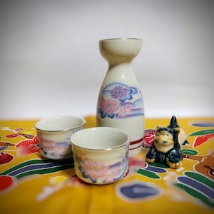 以下が含まれることがあります： 花柄の陶器製の酒器セット。盃3つと徳利1つ、そして小さな猫の置物も付いています。盃と徳利は白地にピンクと紫の花と青いアクセントが施されています。