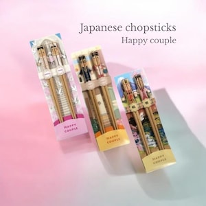 以下が含まれることがあります： 透明なプラスチックパッケージに入った日本の箸の3つのセット。各セットには、カップルを描いた装飾的なトップが付いた木製の箸が1組含まれています。パッケージには「Japanese chopsticks Happy couple」というテキストが含まれています。