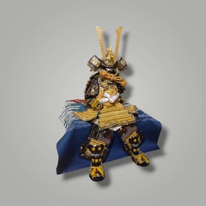 May include: A Japanese samurai doll wearing intricate gold and black armor with a detailed helmet featuring tall golden crests. The figure is seated on a blue fabric base decorated with a white wave pattern, showcasing elaborate metallic textures.