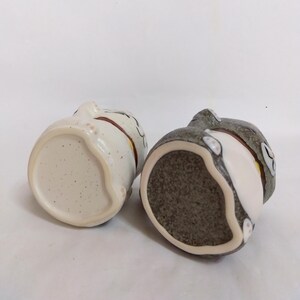 May include: Two ceramic cat figurines, one white with brown and pink accents and the other gray with white and brown accents. Both figurines are shown from the bottom, revealing the unglazed ceramic base.