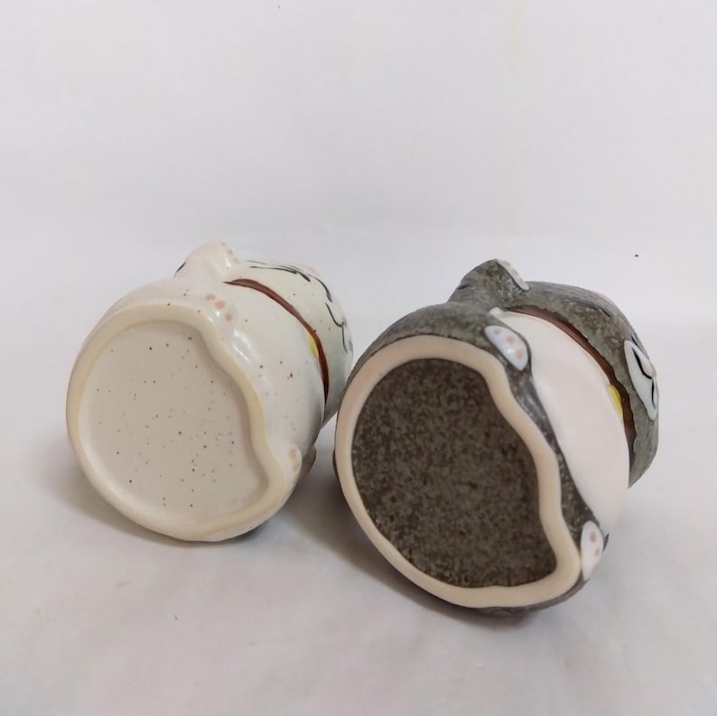 May include: Two ceramic cat figurines, one white with brown and pink accents and the other gray with white and brown accents. Both figurines are shown from the bottom, revealing the unglazed ceramic base.