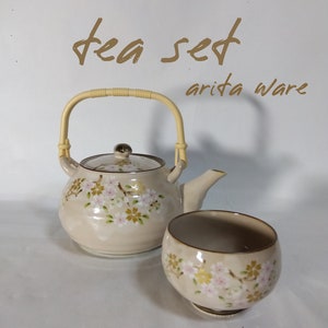 Op de afbeelding: Een beige theeset met een bloemenpatroon. De theepot heeft een bamboe handvat en de kop heeft een bloemenpatroon. De tekst "tea set" en "arita ware" staat op de witte achtergrond.