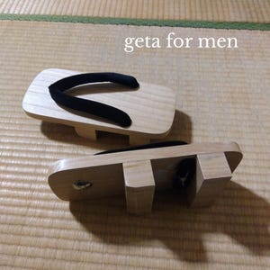 以下が含まれることがあります： 黒い鼻緒が付いた、日本の伝統的な木製下駄2足。サンダルは明るい色の木材で、長方形の台と2つの支持ブロックがあります。「geta for men」というテキストが表示されています。