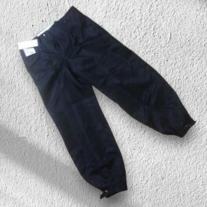 May include: Dark navy blue trousers with a relaxed fit. The trousers have a drawstring waist and a tag attached. The legs taper towards the ankles, which have adjustable closures. The fabric appears to be a lightweight material.