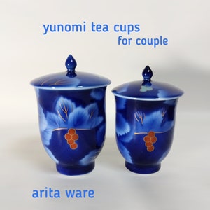 以下が含まれることがあります： ぶどうのデザインが施された、ふた付きの青と白のゆのみ茶碗が2つ。茶碗は有田焼でできており、「yunomi tea cups for couple」とラベルが貼られています。