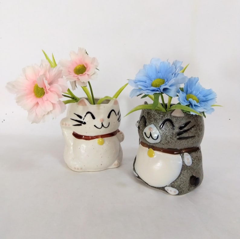 May include: Two ceramic cat planters with smiling faces. One planter is white with pink flowers and the other is gray with blue flowers.