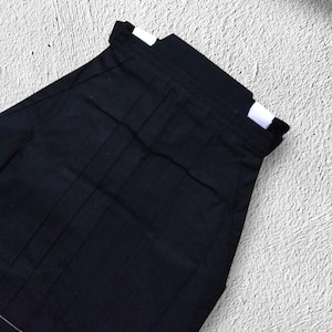 May include: Black hakama pants, a traditional Japanese garment, are displayed against a textured white background. The pants have a pleated design and a wide waistband with white accents. The fabric appears to be a dark, solid color.