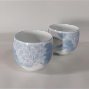 Può includere: Due tazze da tè in ceramica bianca con motivi floreali blu e viola. Le tazze sono rotonde e hanno un leggero bordo.