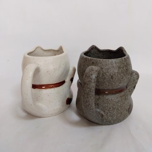 May include: Two ceramic cat mugs, one white with brown speckles and one gray with brown speckles. Both mugs have brown ears, tails, and a brown band around the middle.