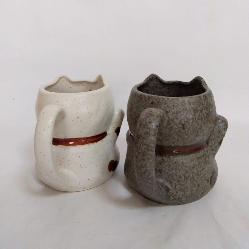 May include: Two ceramic cat mugs, one white with brown speckles and one gray with brown speckles. Both mugs have brown ears, tails, and a brown band around the middle.