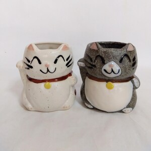 May include: Two ceramic cat planters, one white with brown accents and one gray with brown accents. Both cats are smiling and have their right paw raised in a beckoning gesture.