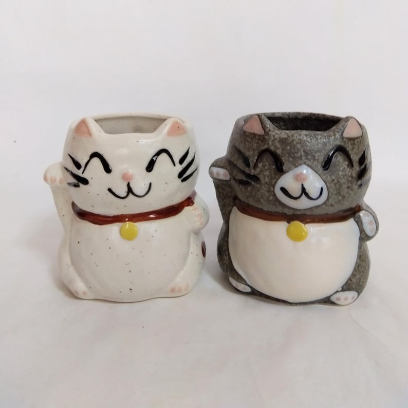 May include: Two ceramic cat planters, one white with brown accents and one gray with brown accents. Both cats are smiling and have their right paw raised in a beckoning gesture.