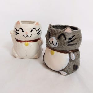 May include: Two ceramic cat planters, one white with brown accents and one gray with brown accents. Both cats have smiling faces and are holding up a paw.