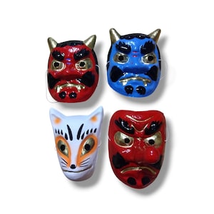 Op de afbeelding: Vier decoratieve maskers gerangschikt in een raster. Linksboven een rood masker met hoorns, rechtsboven een blauw masker met hoorns, linksonder een wit vossenmasker met oranje accenten en rechtsonder een rood masker met een lange neus.