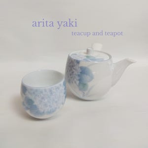 Op de afbeelding: Een witte keramische theepot en bijpassend theekopje met een delicaat blauw en lichtpaars hortensia-patroon. De tekst 'arita yaki' en 'teacup and teapot' is in het lichtpaars boven de objecten op een effen witte achtergrond gedrukt.