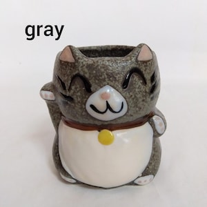 May include: Gray ceramic cat planter with a white belly, brown collar, and a yellow bell. The cat has a happy expression with black eyes and a pink nose.